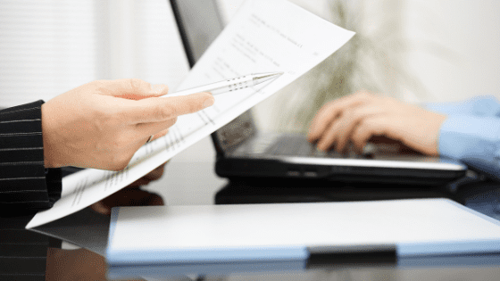 a person with a document in his hand- another person typing on the laptop